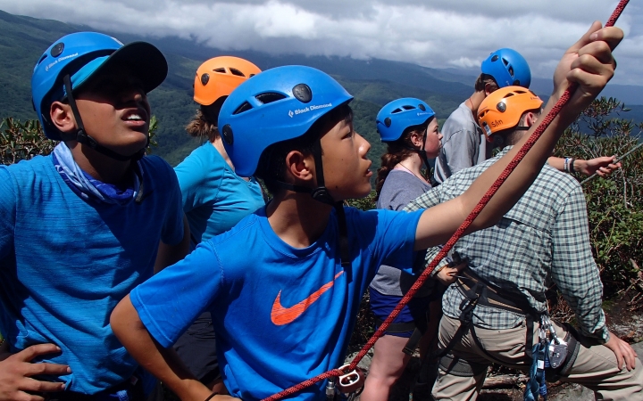 Students wearing helmets and harnesses belay rock climbers, while others spot them. They appear to be at high elevation, above a green forrest. 